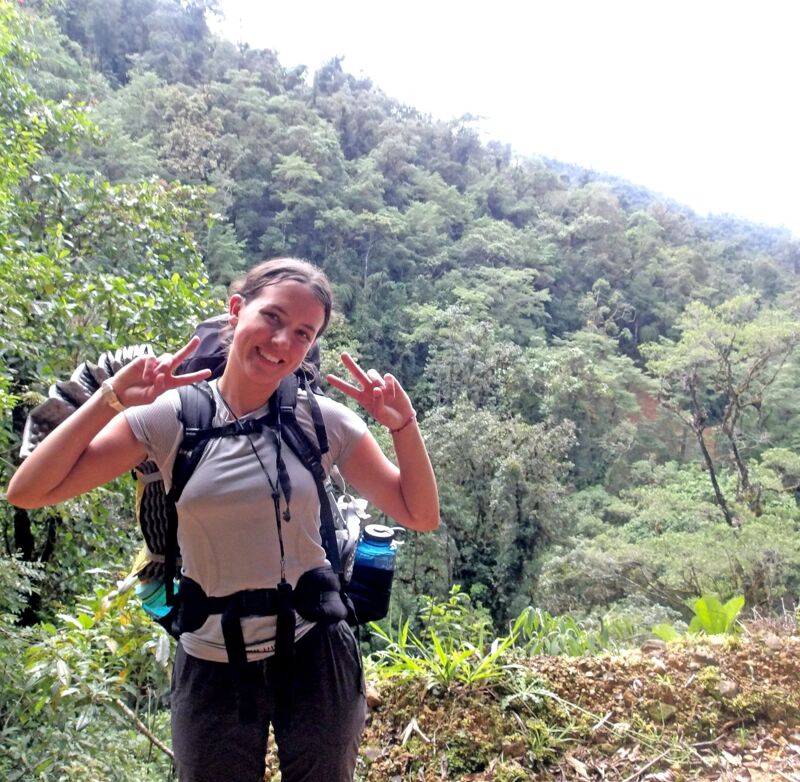 A woman with a backpack is standing in front of a lush green forest. She is wearing a grey t-shirt and dark pants. She is holding up two peace signs with both hands and smiling. The background is filled with dense trees and vegetation, suggesting a natural setting, possibly a hiking trail or forest path.
